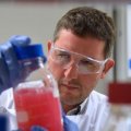 A male scientist in lab coat and goggles inspects a glass full of red liquid. 